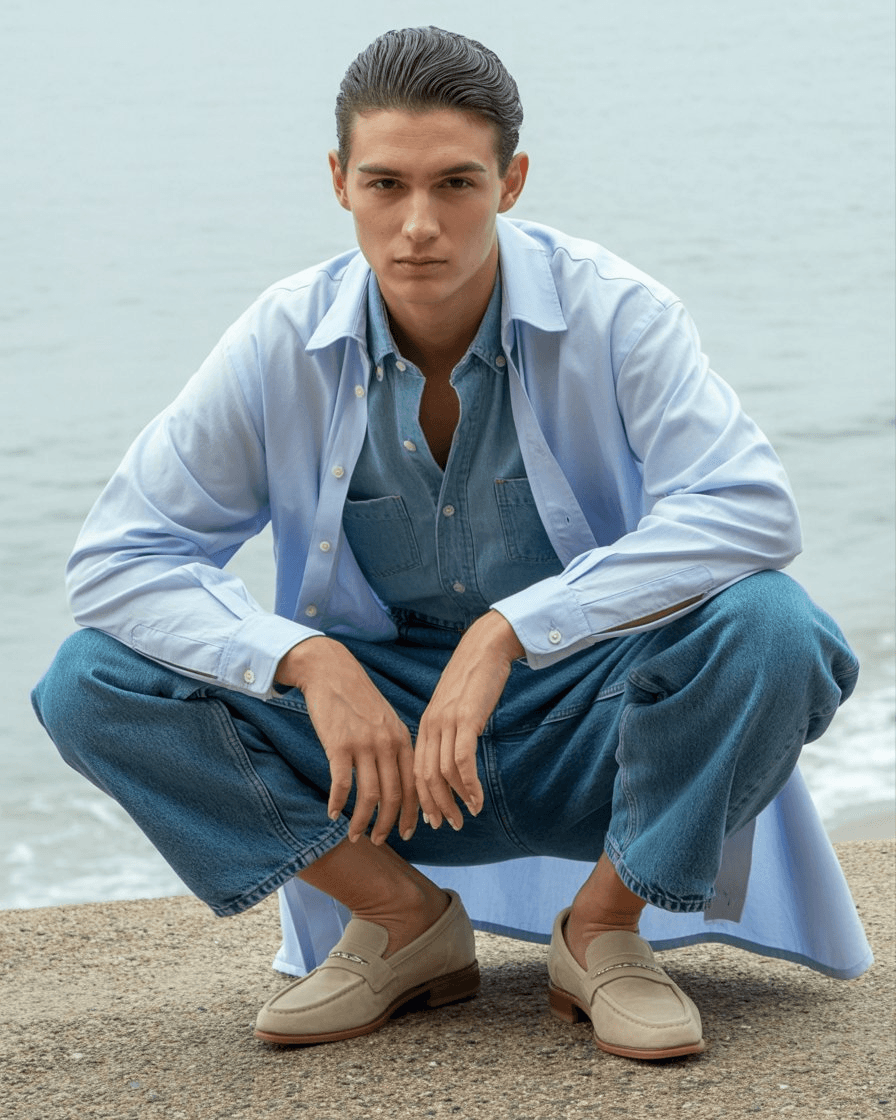 Oversized Pale-Blue Poplin Shirt Over A Chambray Shirt, Paired With Relaxed Blue Jeans And Beige Suede Loafers, Emphasizing A Loose And Effortless Silhouette