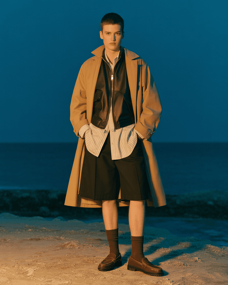 Khaki Trench Coat Layered Over White Shirt And Black Leather Zip Vest, Matched With Black Knee-Length Shorts, Brown Socks, And Brown Loafers For A Utilitarian Look